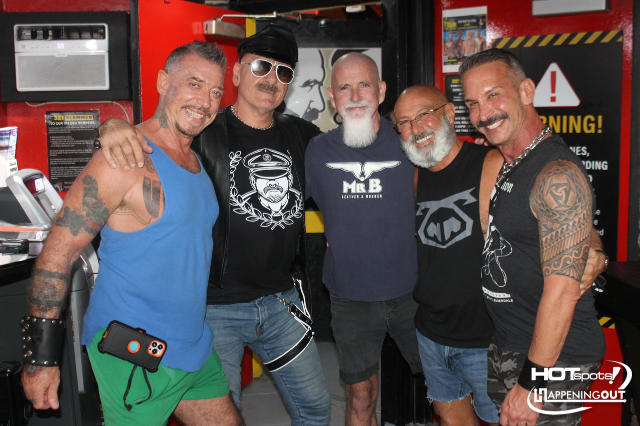 Group of five men posing together in a colorful, edgy club setting; everyone smiles and arms around each other, showcasing tattoos and casual rock style.—informative image