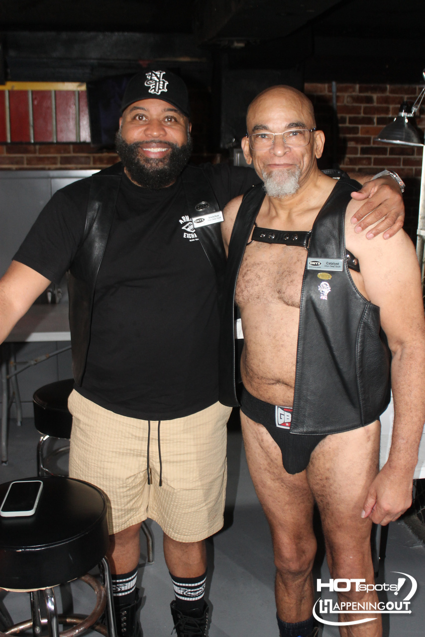 Two men pose together in a backstage-like area; one wears a black cap and t-shirt, the other a leather vest and briefs, smiling at the camera.