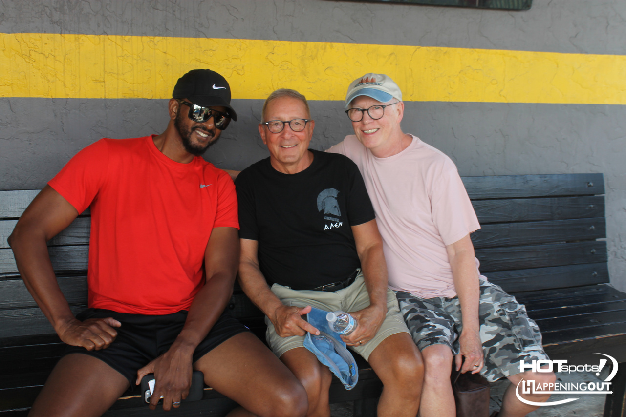 Three men sit on a black bench against a gray wall with a yellow stripe, smiling at the camera.