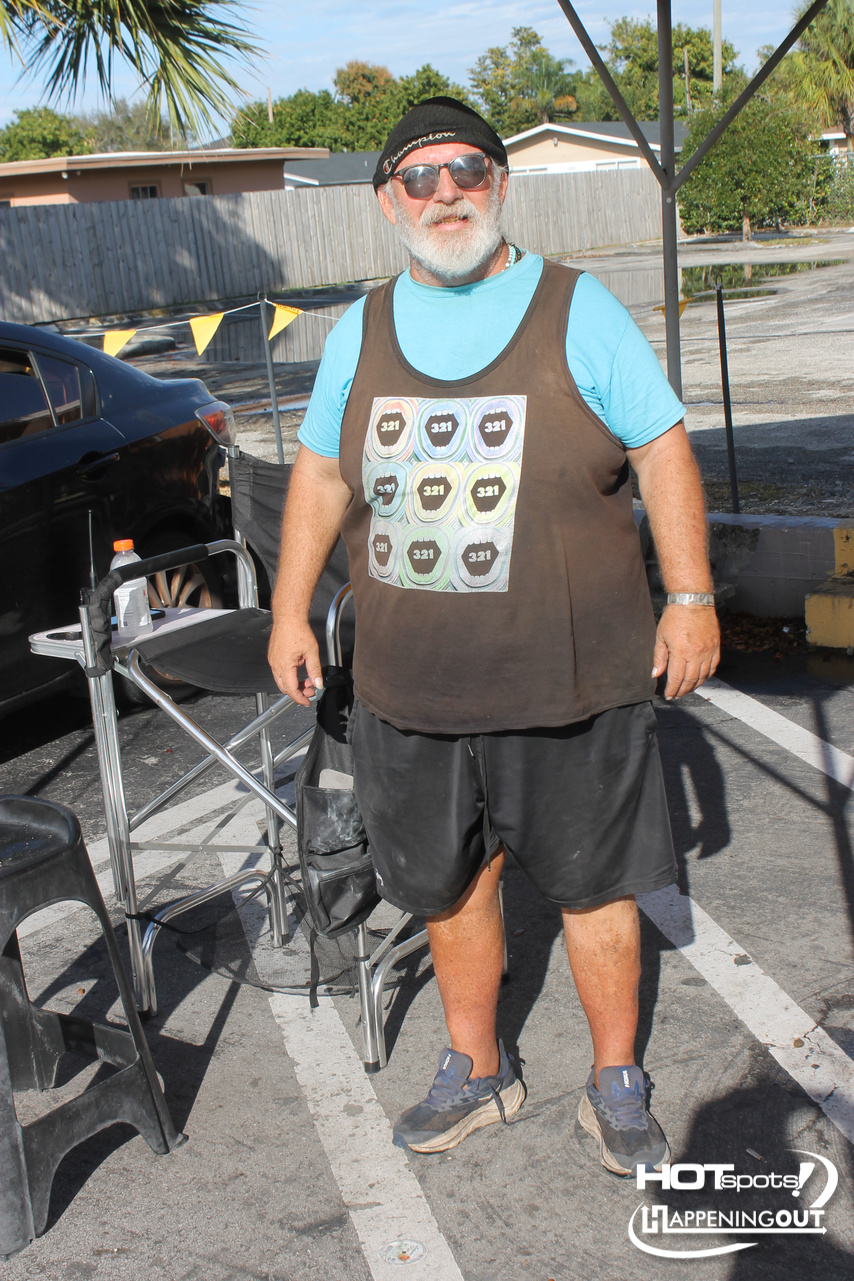 Older man with white beard and sunglasses, wearing a teal shirt under a brown sleeveless top, black shorts, and sneakers, standing outdoors at a casual event by a car and folding chair.