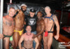 Six muscular men pose together in a club backstage area, smiling and with arms around each other after an event.