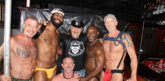 Six muscular men pose together in a club backstage area, smiling and with arms around each other after an event.