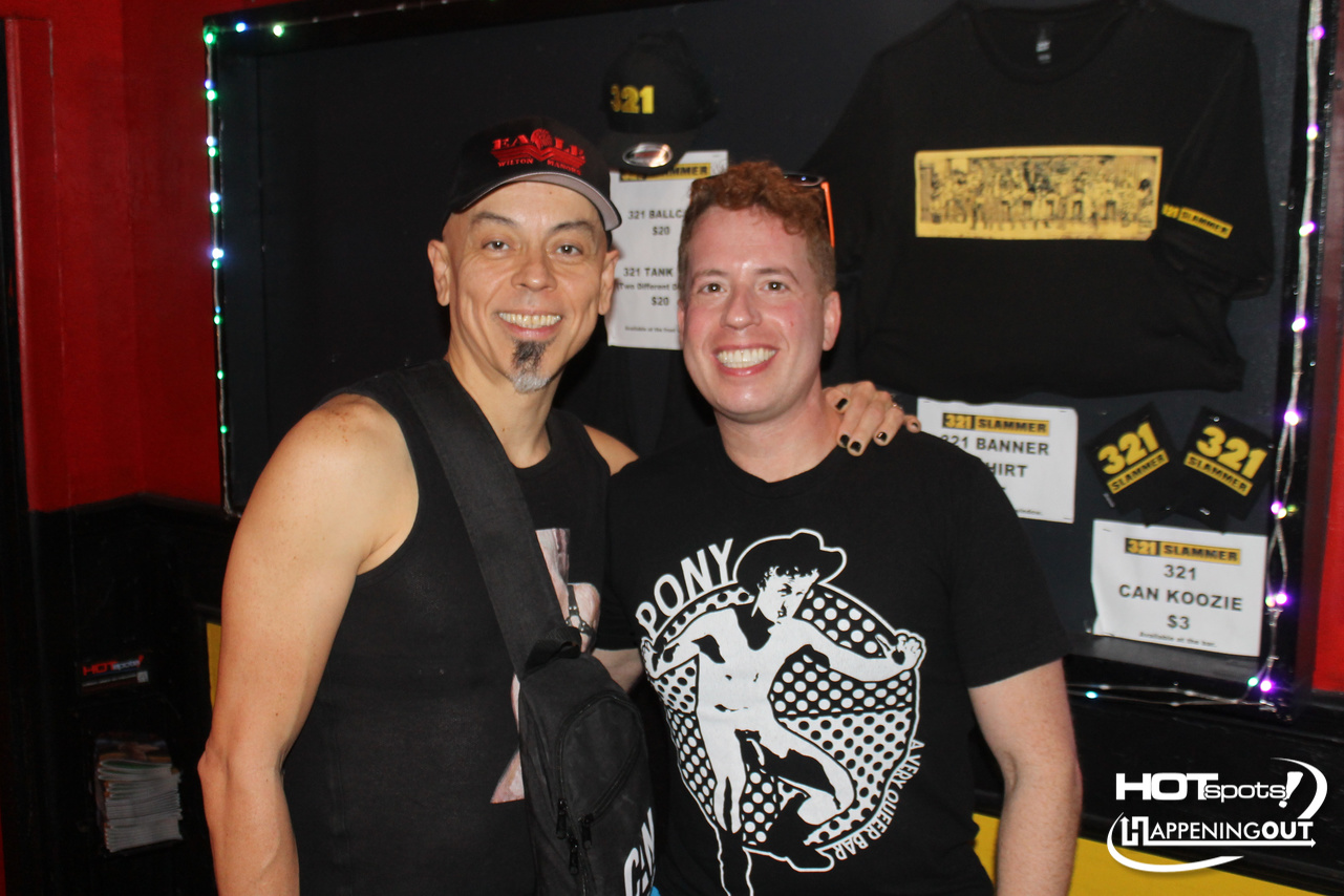 Two smiling men posing together at an event, with merchandise shirts on display in the background and colorful lights.