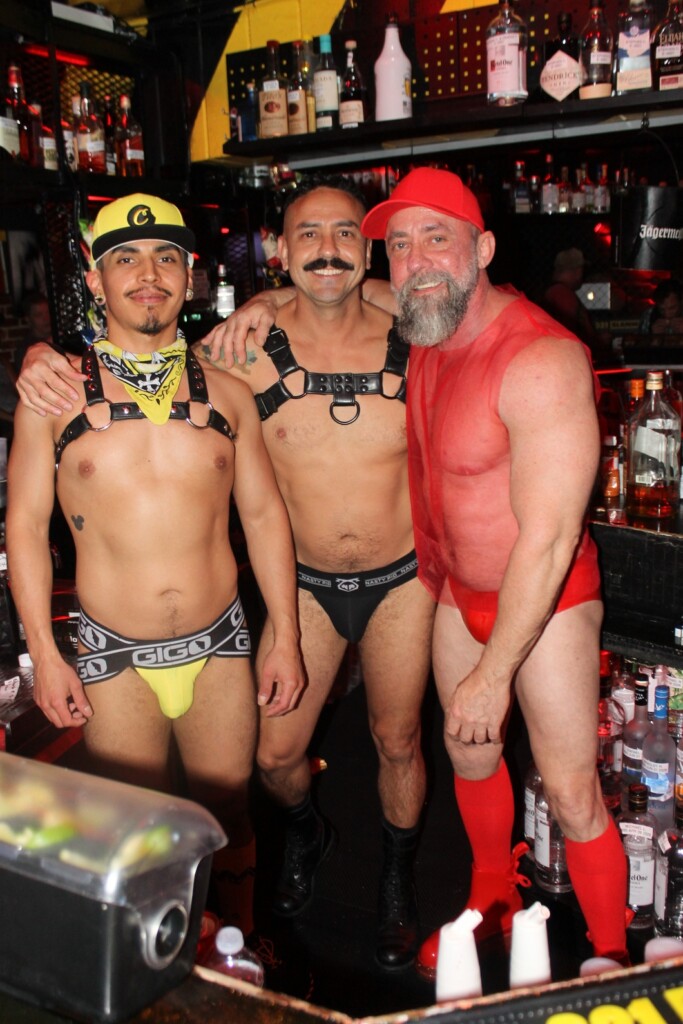Three men pose behind a bar in fetish gear: two shirtless with leather harnesses and briefs, one in a red mesh shirt and red cap.