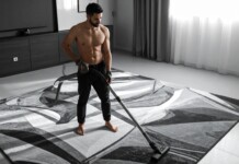 Shirtless man vacuuming a large patterned rug in a modern living room.