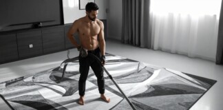 Shirtless man vacuuming a large patterned rug in a modern living room.