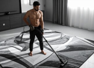 Shirtless man vacuuming a large patterned rug in a modern living room.