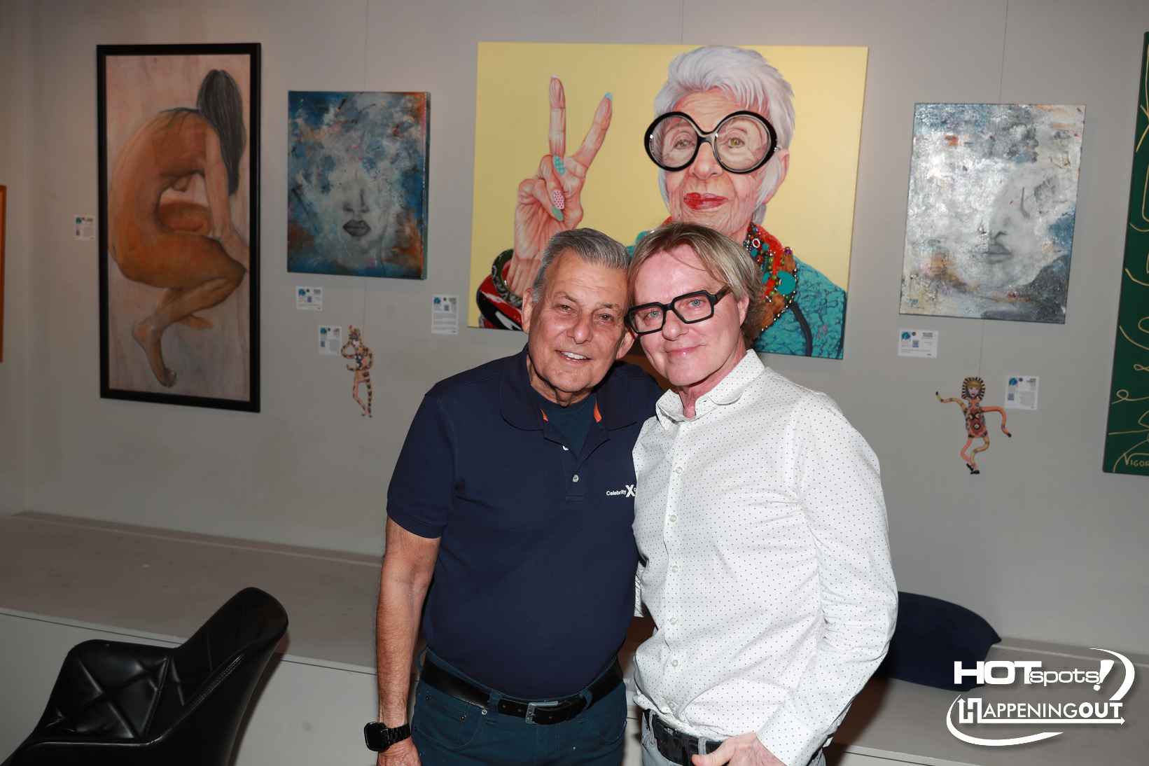 Two men pose together in an art gallery, smiling in front of colorful paintings on the wall.