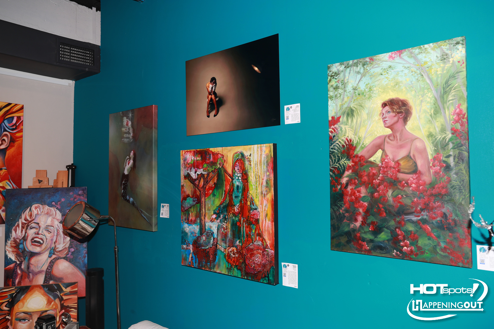 Teal gallery wall with multiple colorful paintings, including a woman in a garden on the right side of the wall.
