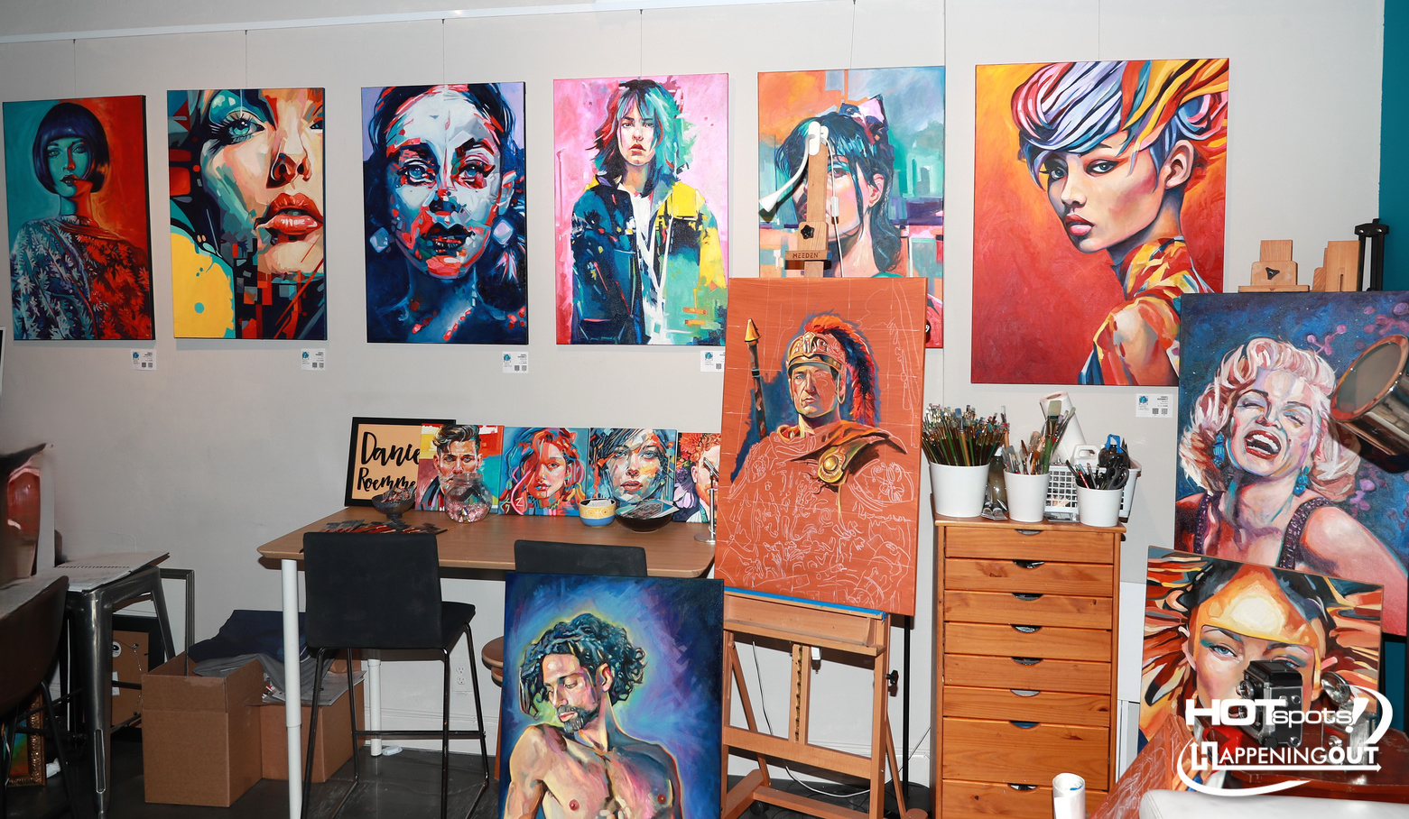 Vibrant art studio with a wall of bold, colorful portrait paintings and an easel with a large orange portrait in the foreground.