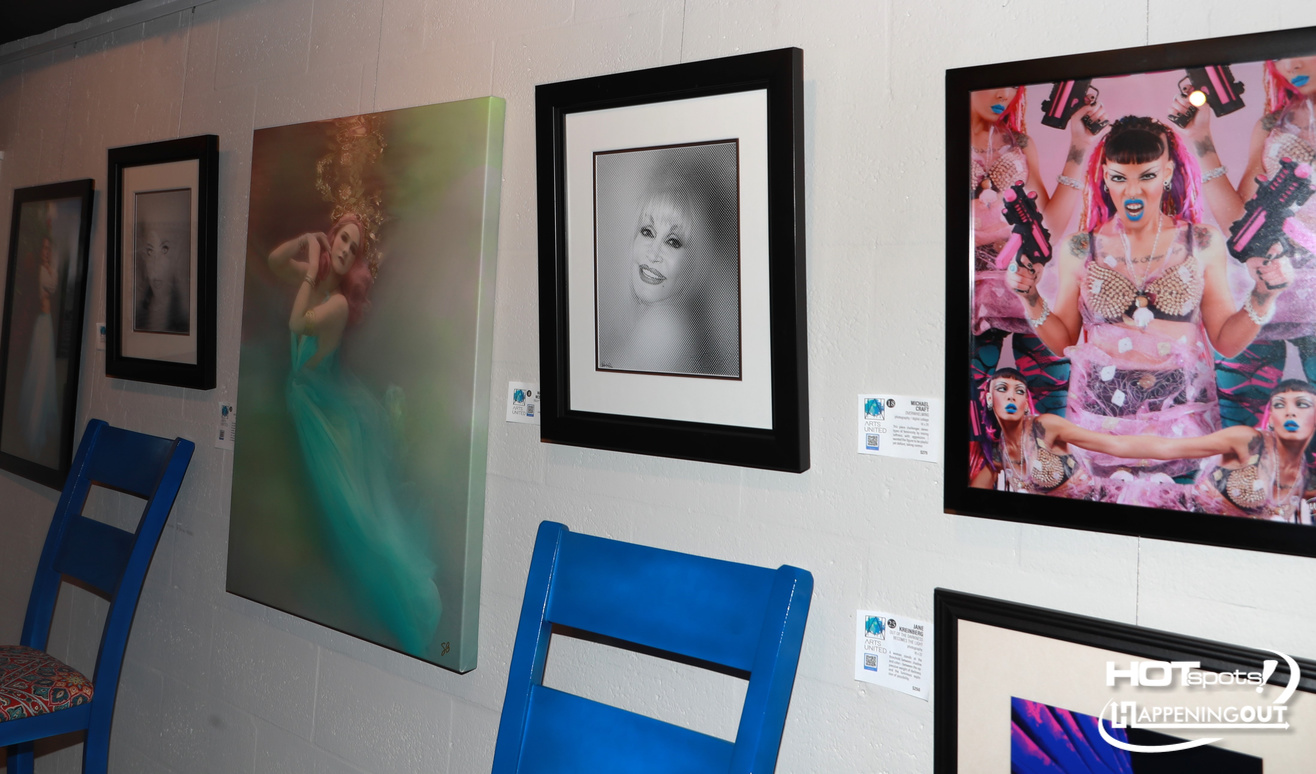 Gallery wall with framed portraits and a teal dress photo, blue chairs in the foreground.