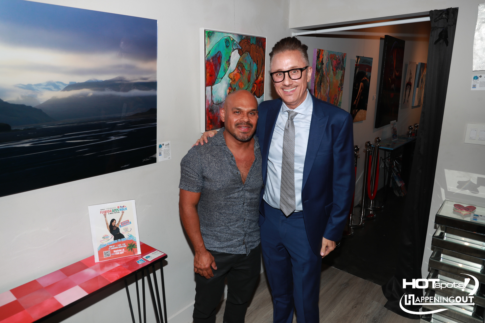 Two men posing together in an art gallery, one in a blue suit with glasses and the other in a patterned grey shirt with short sleeves, smiling at the camera.