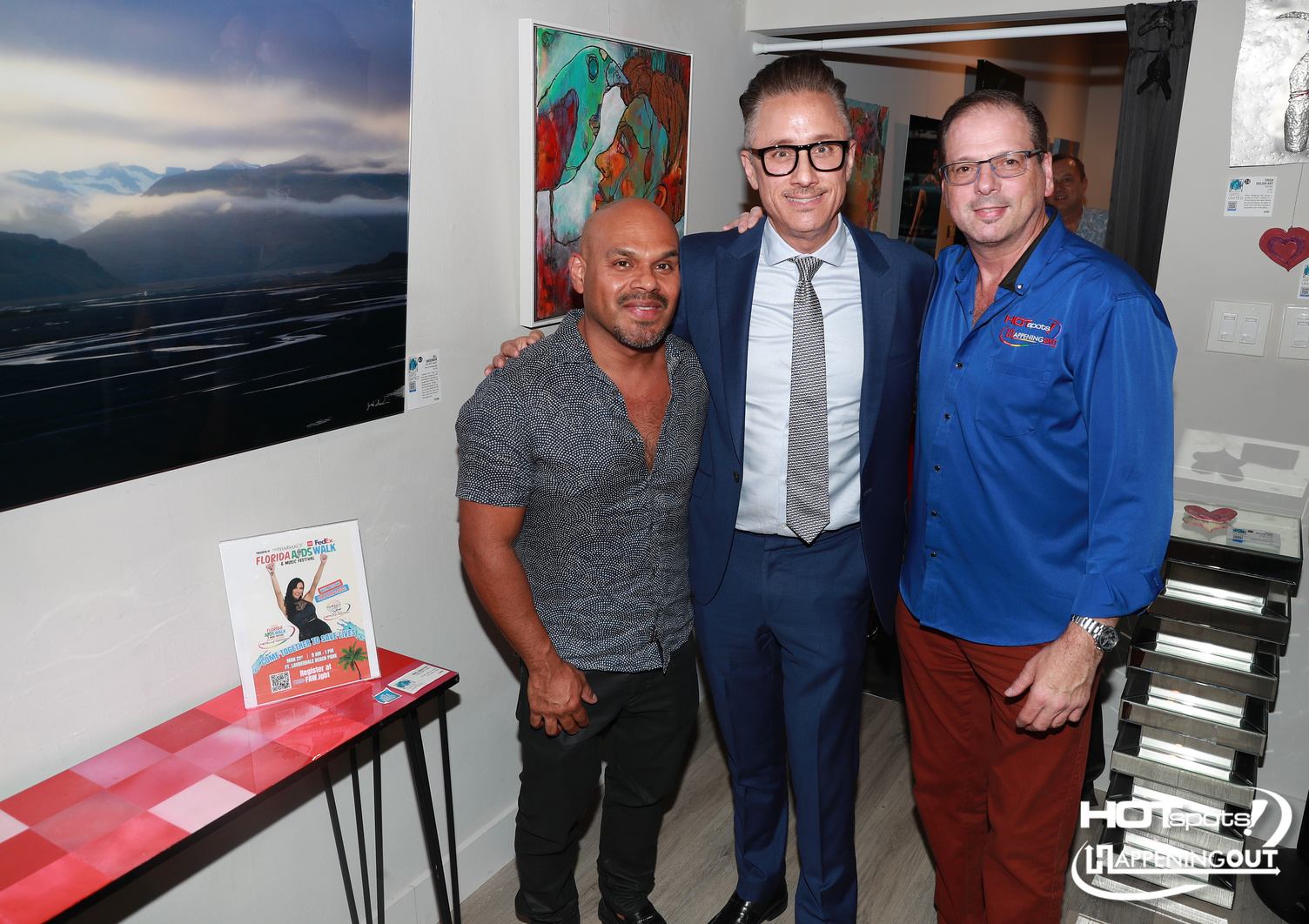 Three men pose together at an art gallery or event, smiling for the camera, with colorful paintings on the wall behind them.