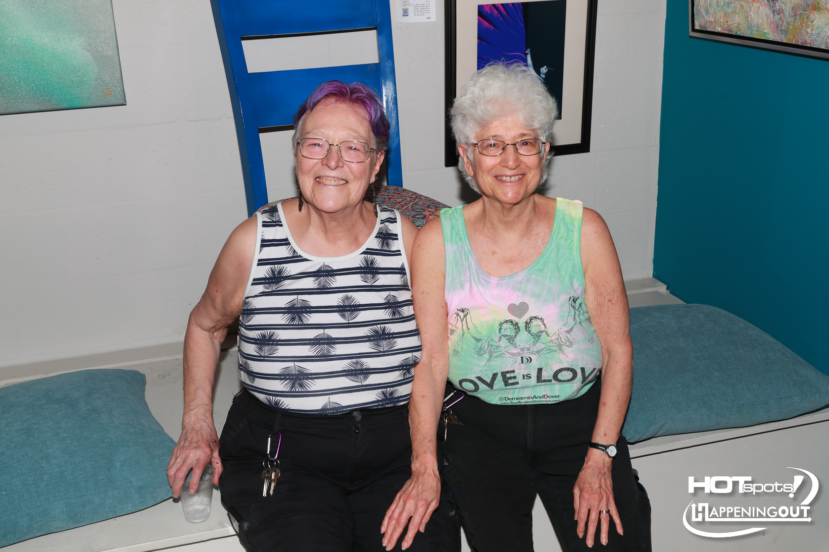 Two smiling older women sit close together on a teal bench in an indoor art gallery, wearing sleeveless tops and glasses.