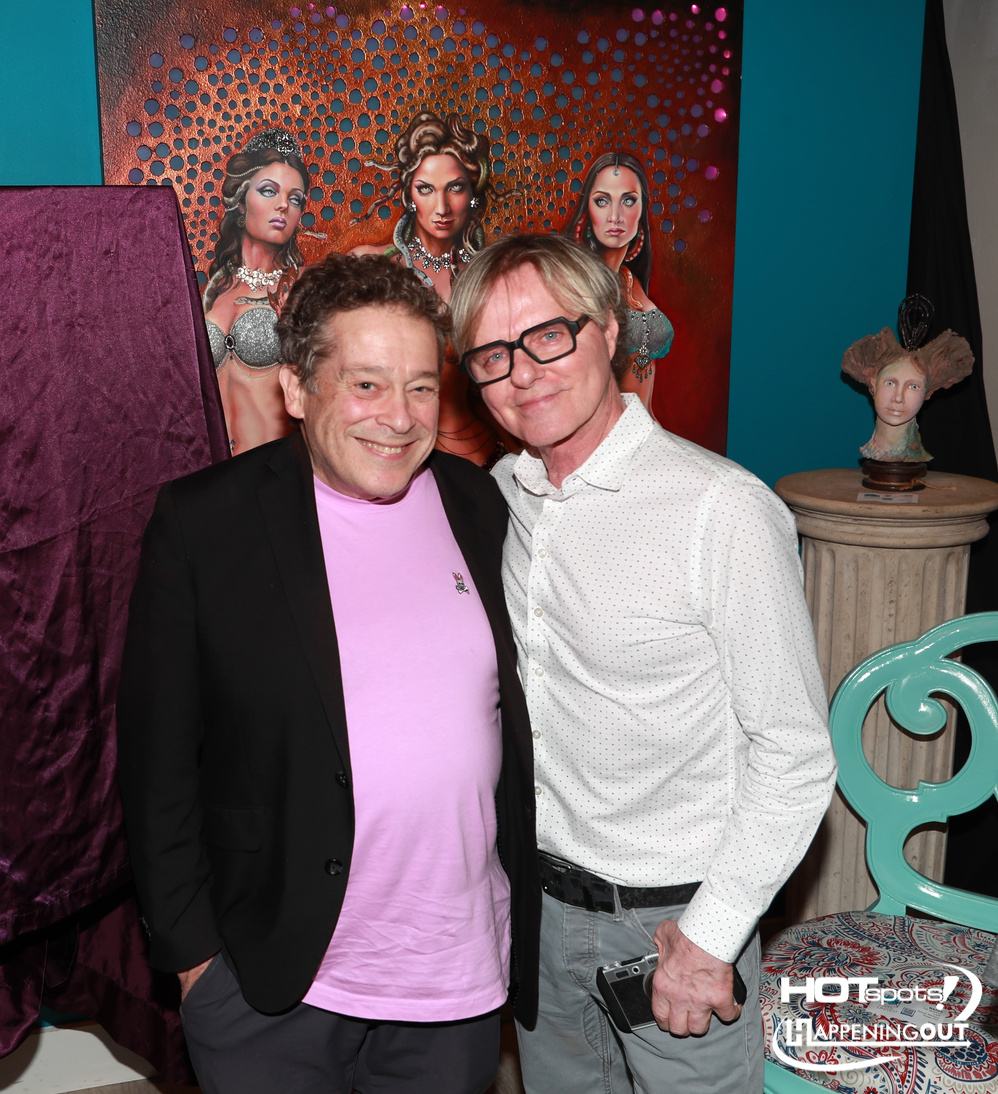 Two men smiling for a photo at an art-themed event, one in a pink shirt and black blazer, the other in a white polka-dot shirt with glasses, in front of a vibrant mural of three women.