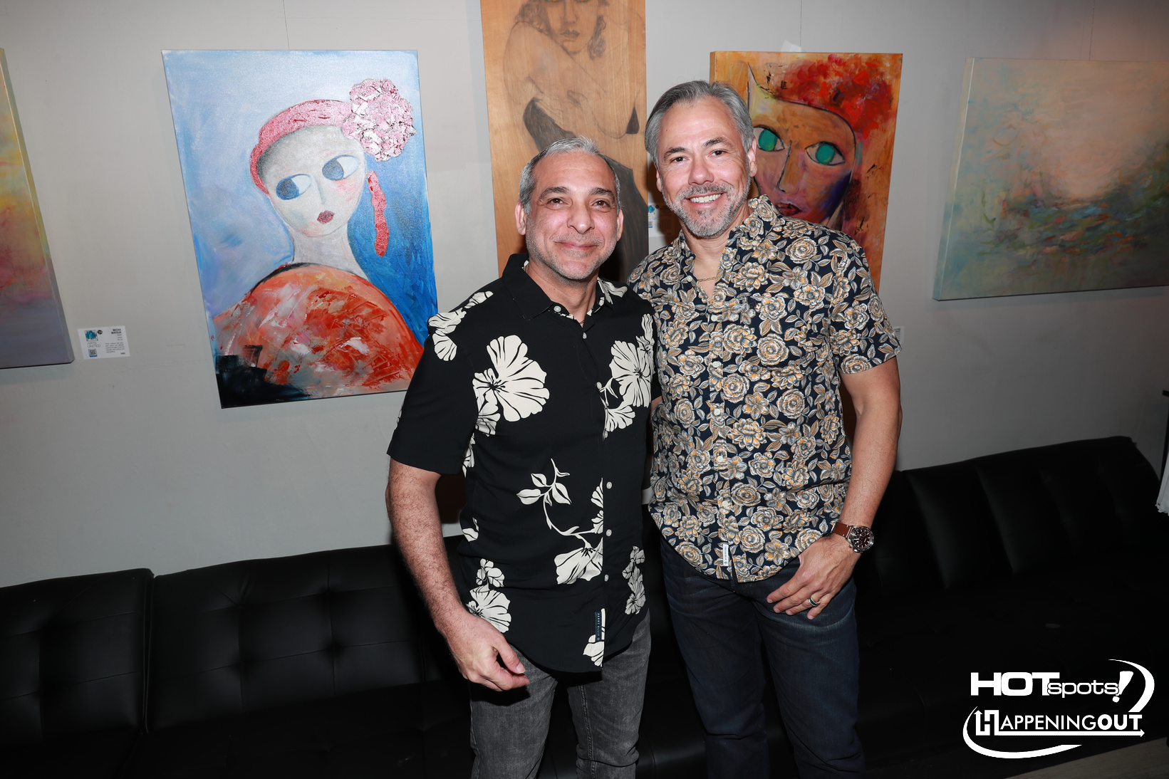 Two smiling men pose together in an art gallery, wearing tropical-pattern shirts, with colorful paintings in the background.