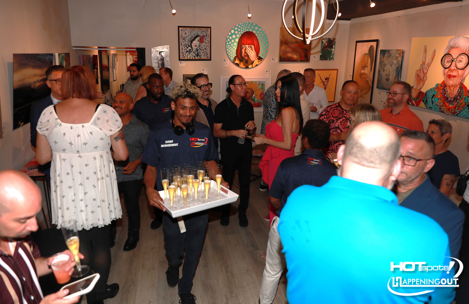 Crowded art gallery reception with guests mingling and a server carrying a tray of champagne flutes in the foreground.