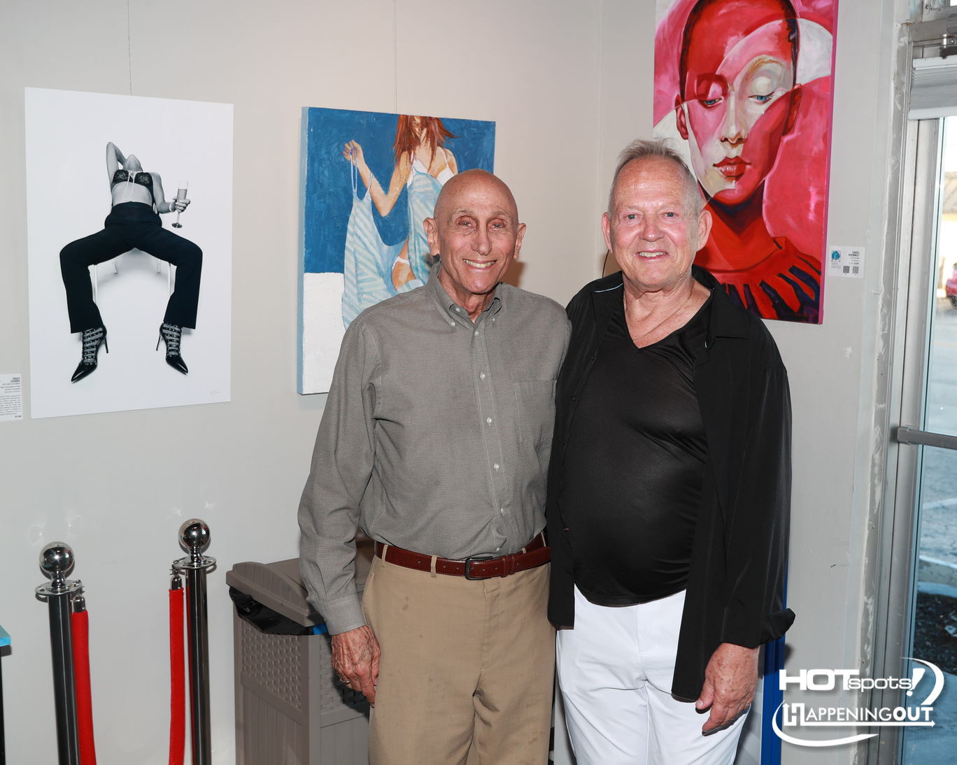 Two smiling men pose in an art gallery with large paintings on the wall behind them.