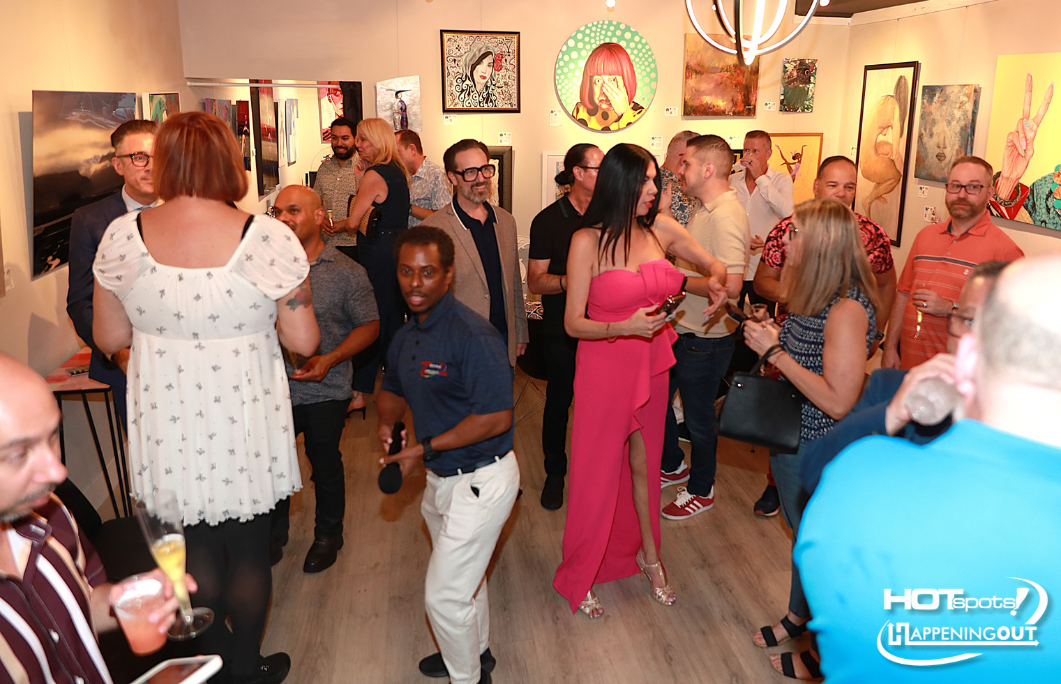 Crowded art gallery opening with guests socializing and admiring framed paintings on walls.