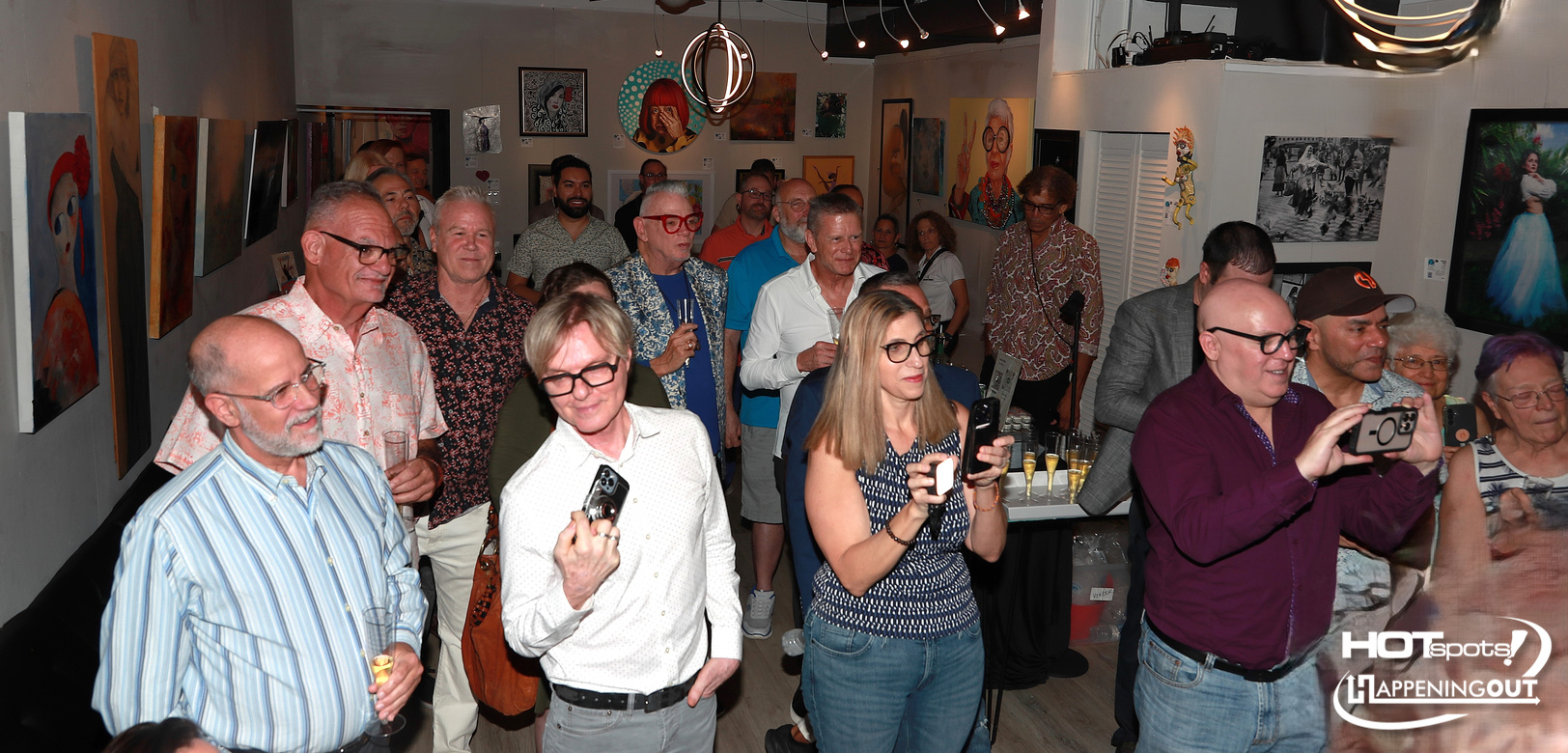 Crowded art gallery reception with guests mingling and photographing framed paintings on the walls.