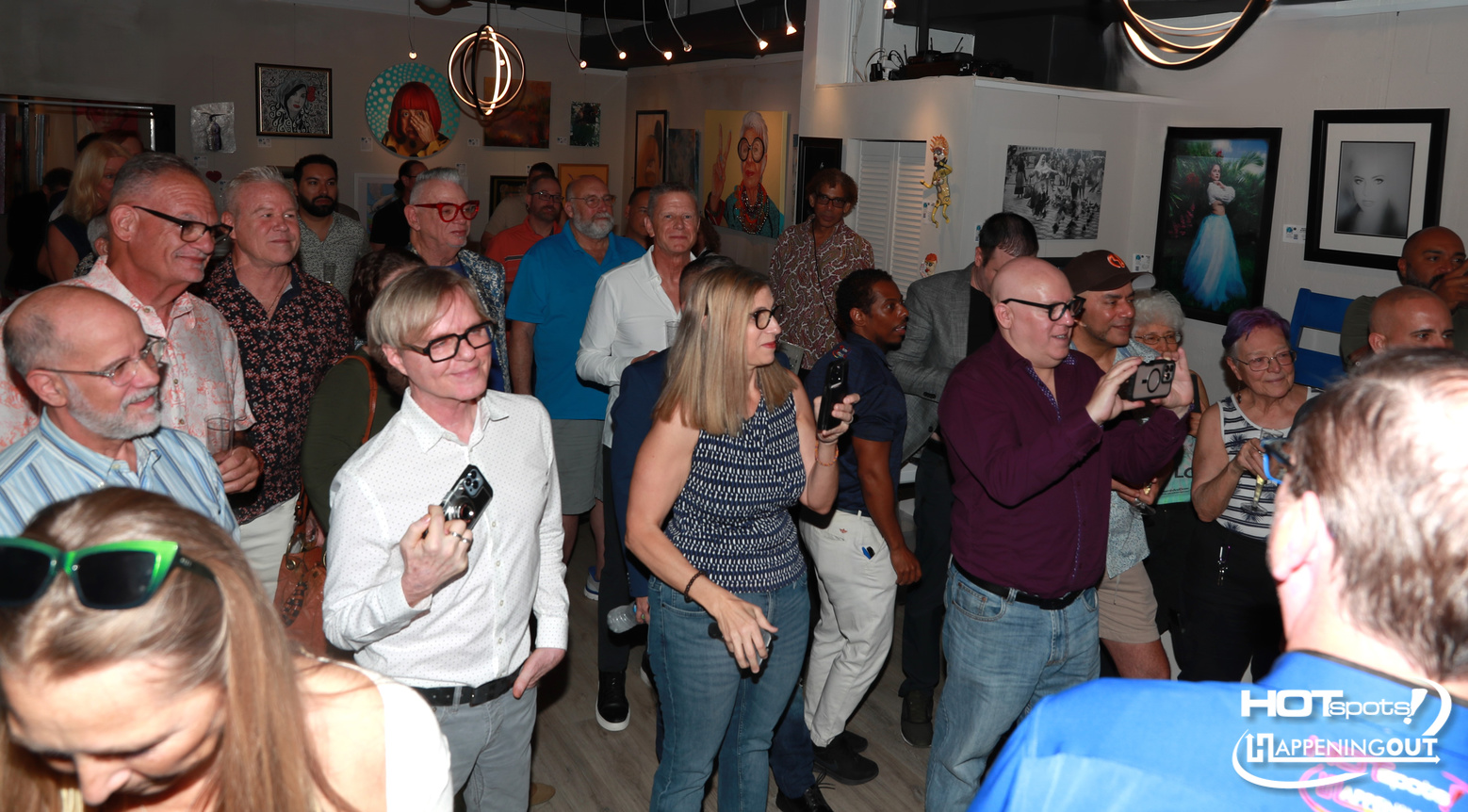Crowd of people at an art gallery event, many holding cameras or phones to take photos of the artwork on the walls.