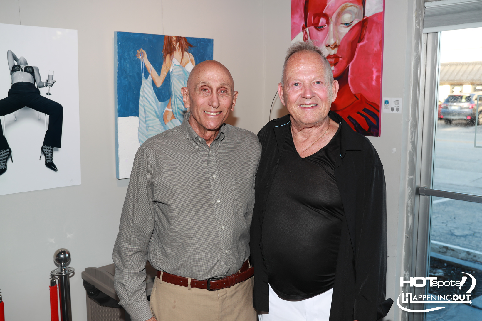 Two smiling older men pose together in an art gallery with colorful paintings on the wall behind them, including a red-pink abstract piece on the right and a blue-toned figure in the middle.