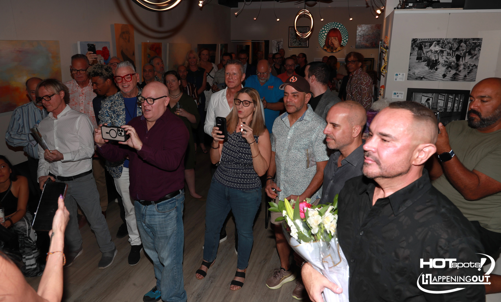 Crowd of people at an art gallery event, many filming or taking photos with smartphones.