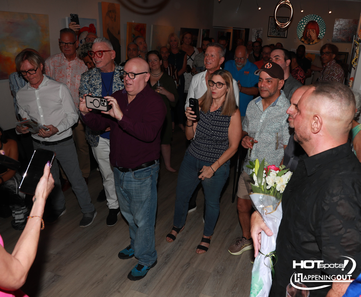People gathered at an art gallery event; many hold phones to record, a man in a maroon shirt films, and a bouquet is visible in the foreground.