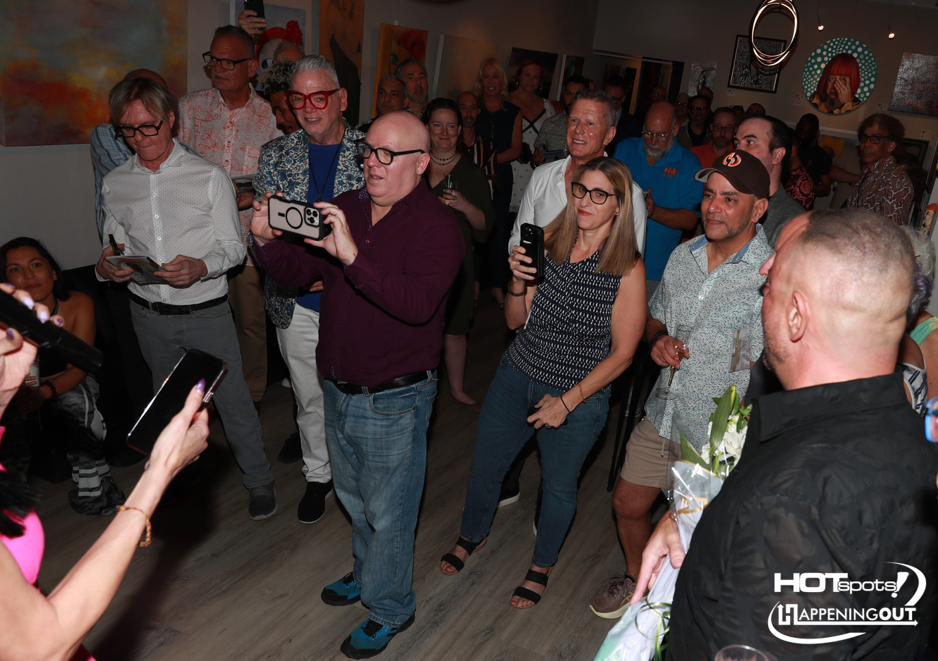 Group of guests at an art gallery event, many holding phones to record the moment, colorful paintings on the walls.