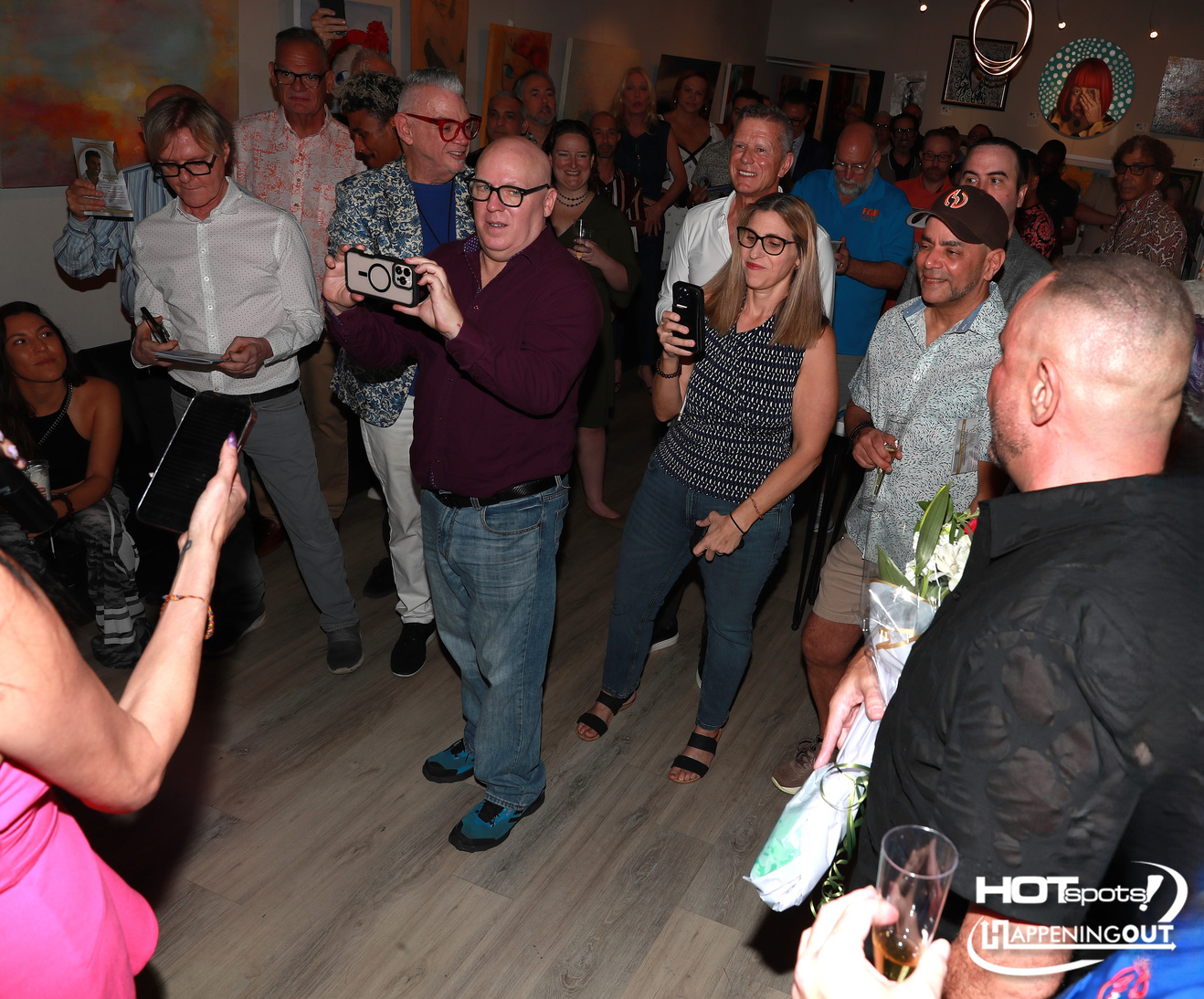 Crowd at an art gallery party, many people holding phones to photograph the moment with colorful paintings on the walls behind them.