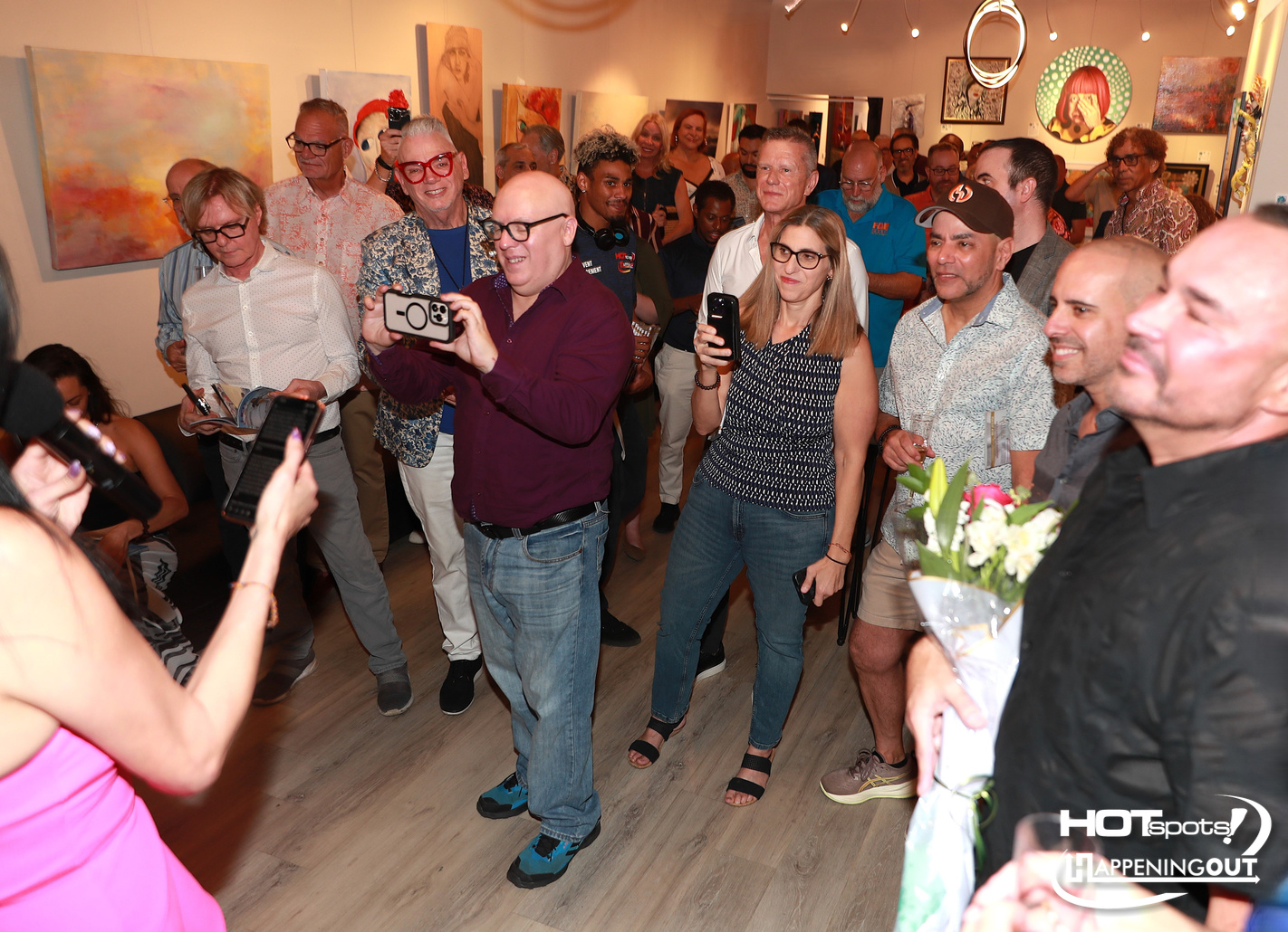 Crowd of people at an art gallery opening, some taking photos with phones and cameras, with colorful paintings on the walls behind them.