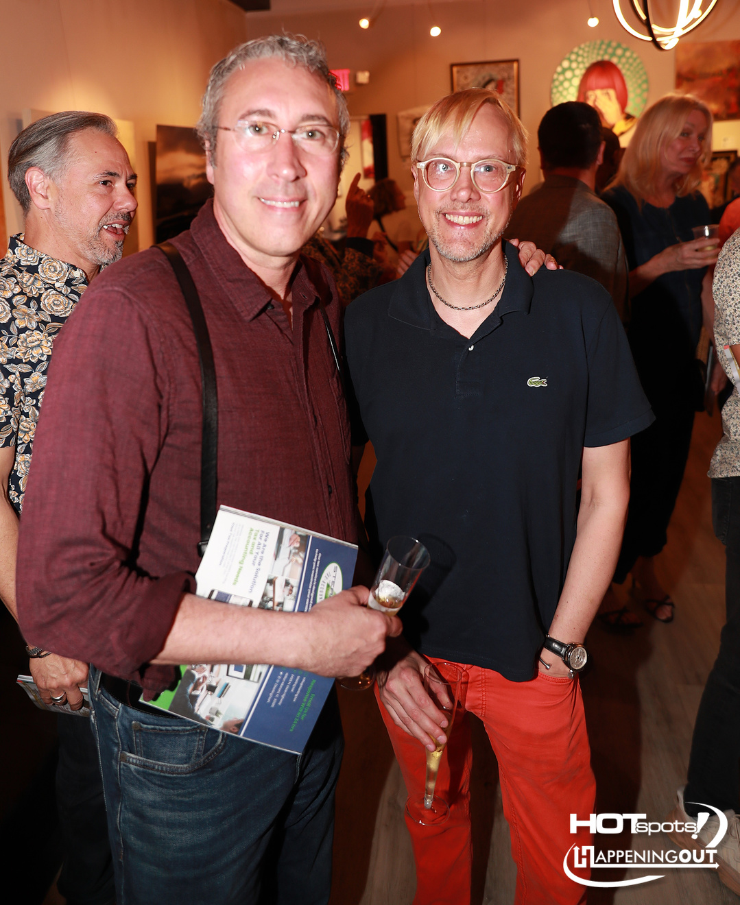 Two men posing at an art gallery reception; one in a maroon shirt holds a brochure and a champagne flute, the other in a navy polo and orange pants smiles beside him.