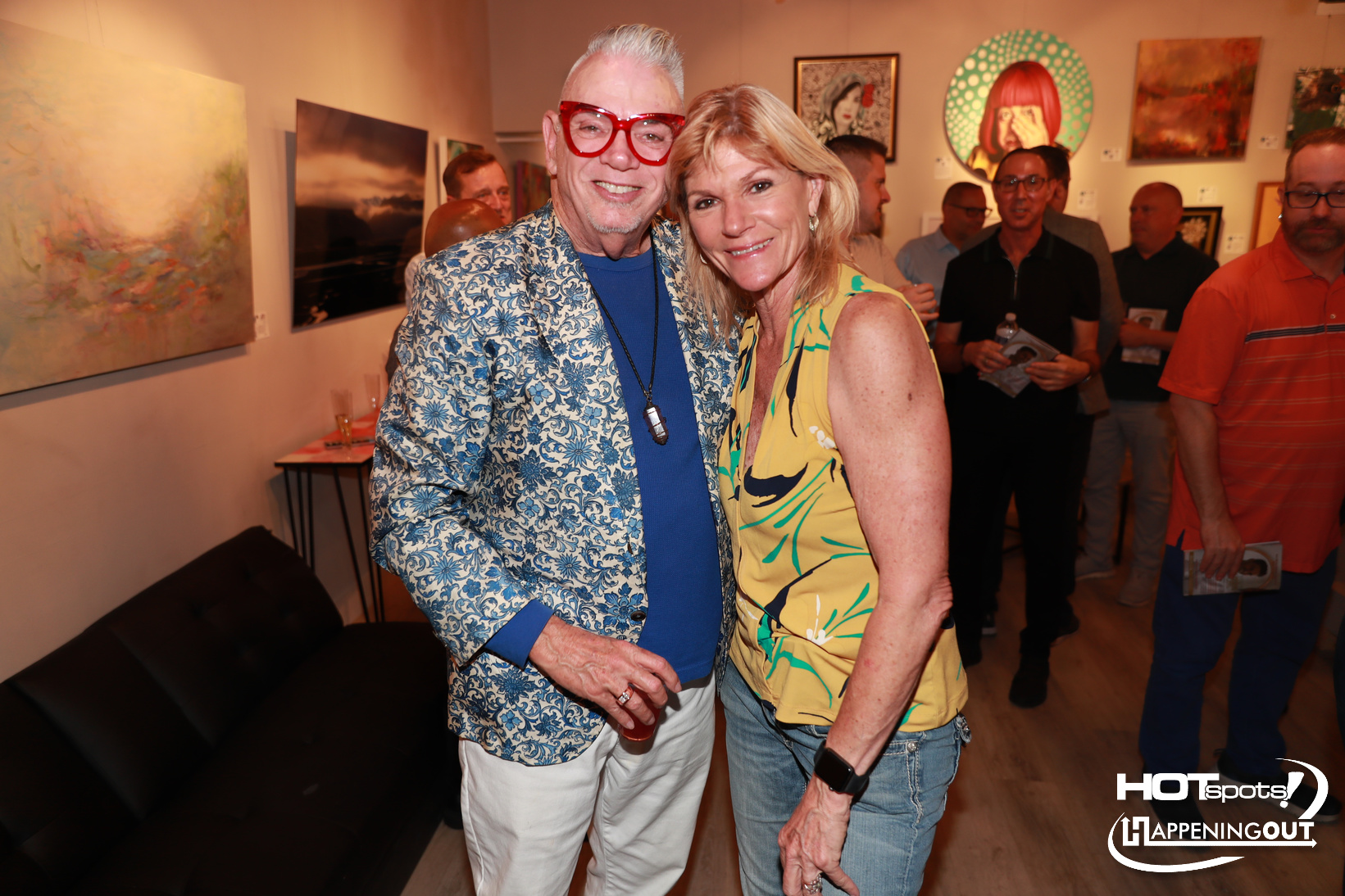 Two smiling attendees pose at an art gallery opening, with paintings on the wall and a group of people in the background.