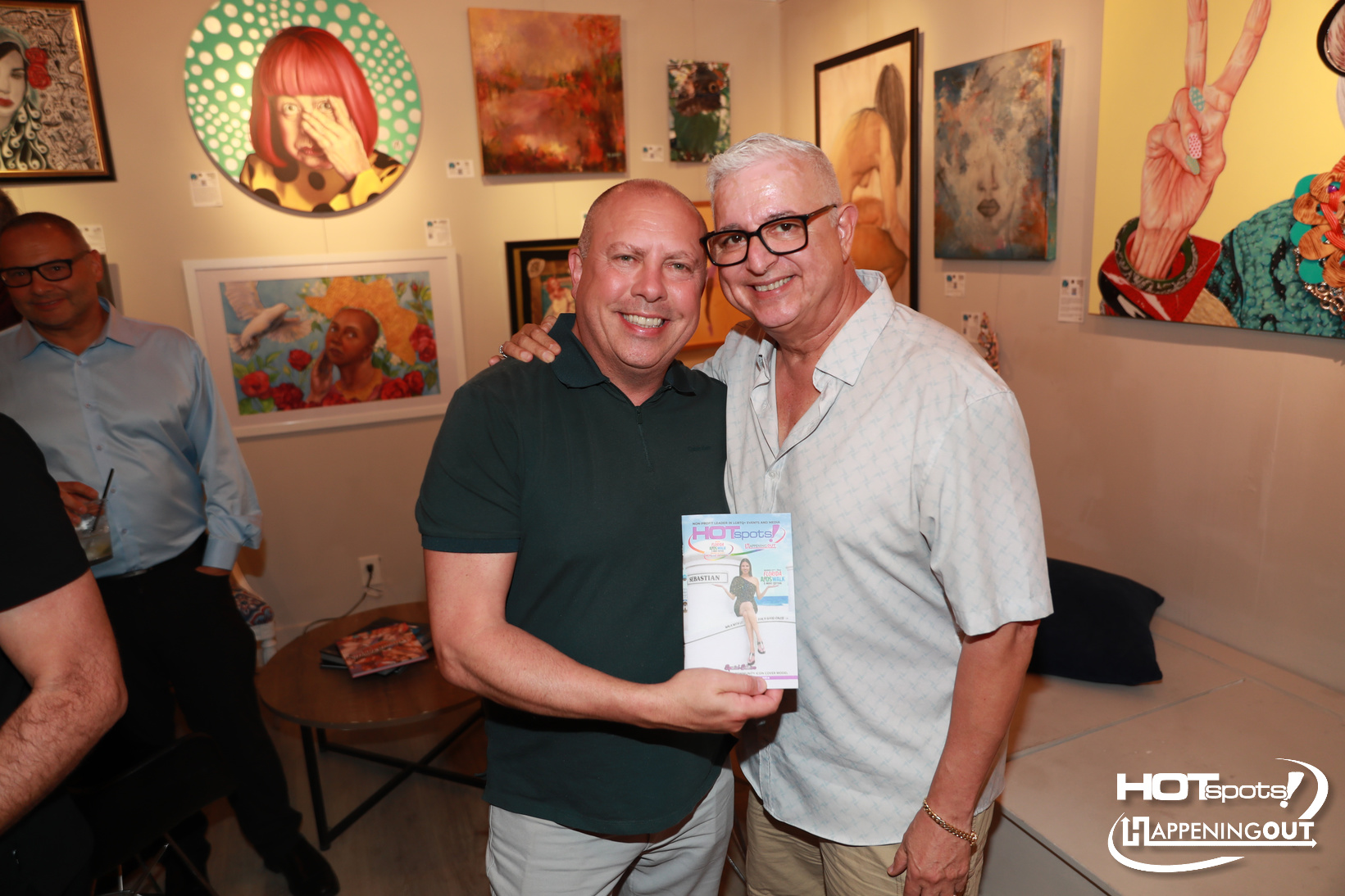 Two men smiling for a photo at an art gallery; one holds a glossy magazine or program in front of them.