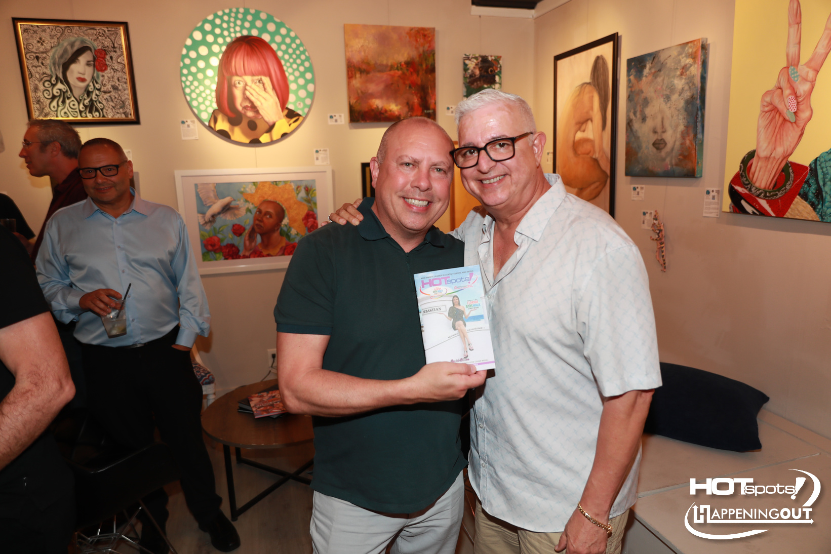 Two men smile at the gallery, one holding a HOTspots magazine as they pose together in front of colorful artwork.