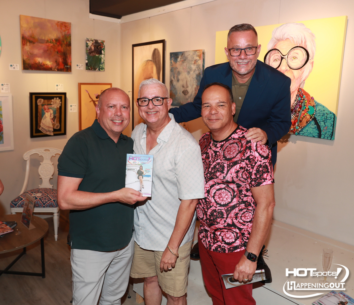 Four men pose together in an art gallery, one holding a magazine, with vibrant paintings on the walls and smiling for the camera.
