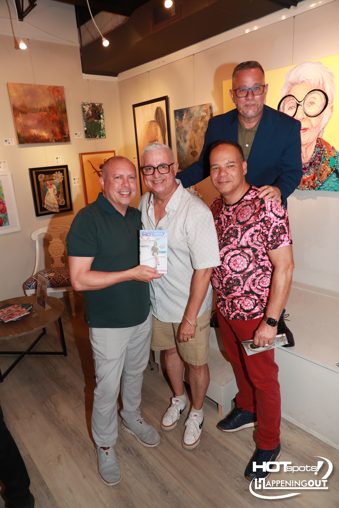 Four men pose together at an art gallery; the man in green polo holds a flyer while others smile, with colorful paintings in the background.