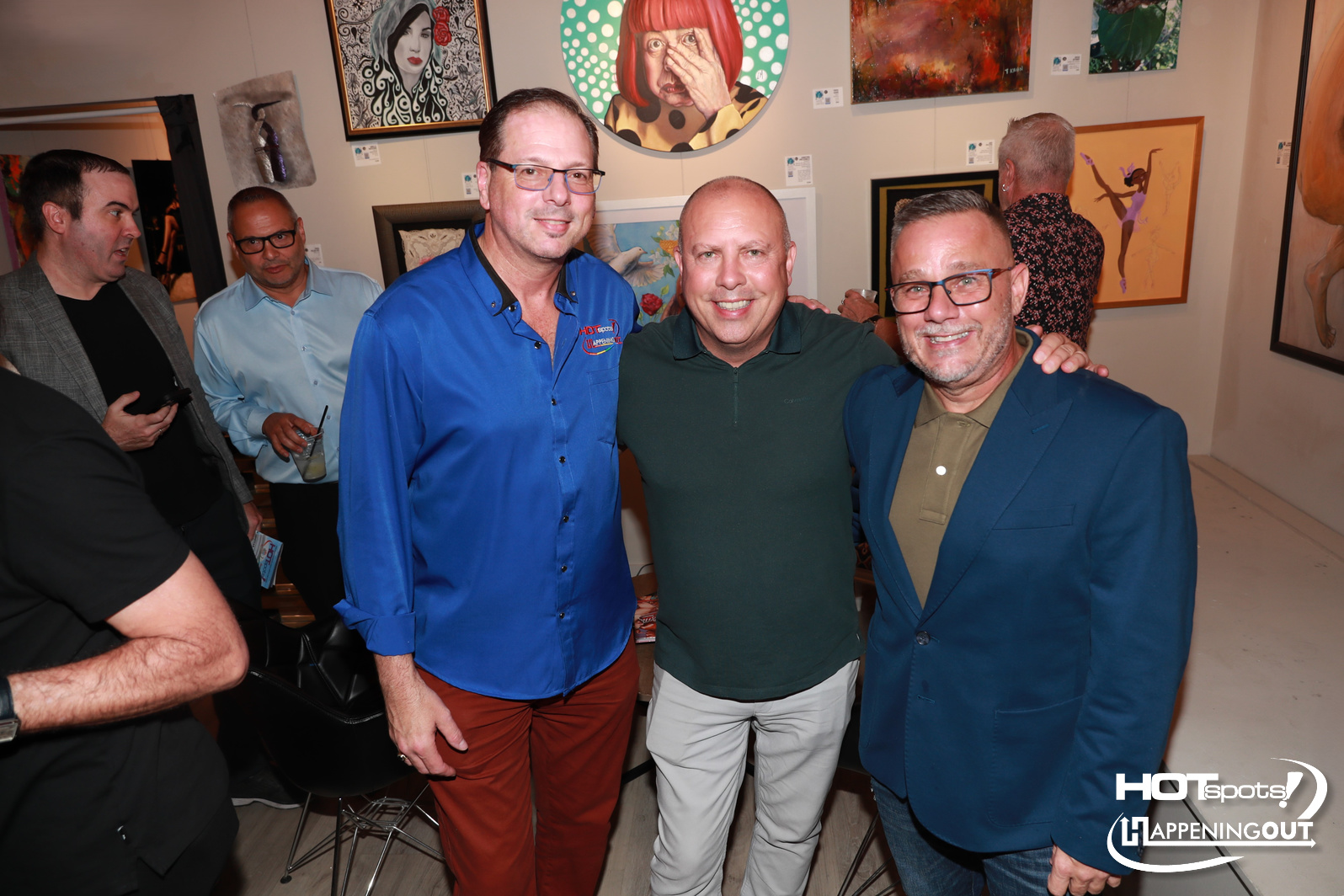 Three men pose for a photo at an art gallery opening, with colorful paintings on the wall behind them and small label cards nearby.