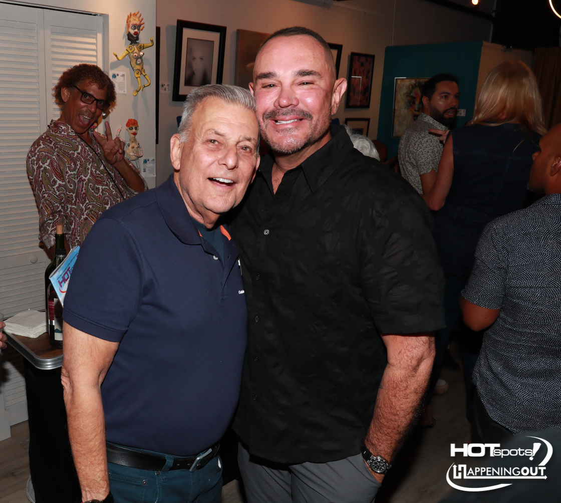 Two men smiling for the camera at an art gallery party, one in a navy polo and the other in a black shirt, with artwork on the wall behind them.