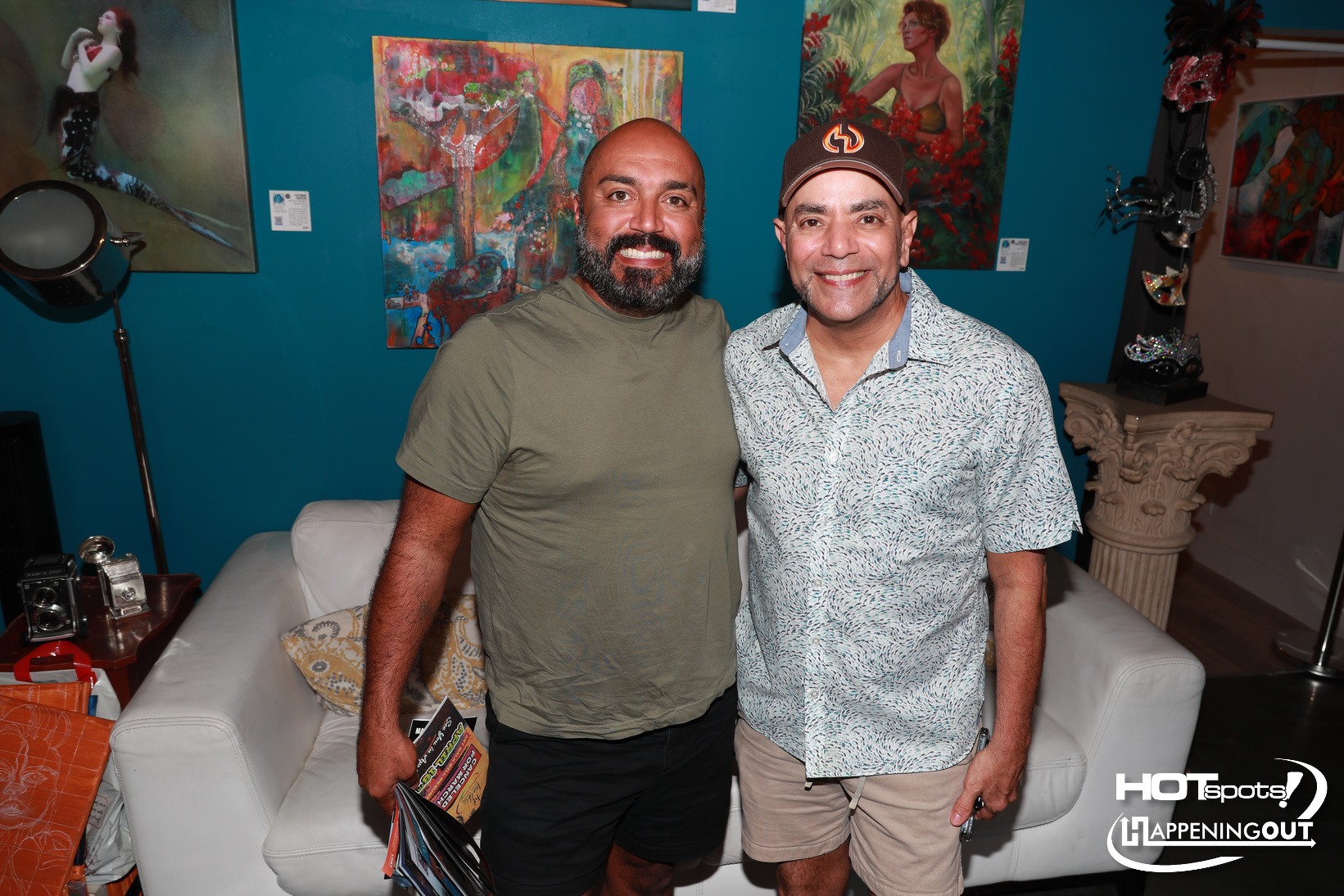 Two men smiling and posing in an art-filled lounge, turquoise walls and colorful paintings behind them.