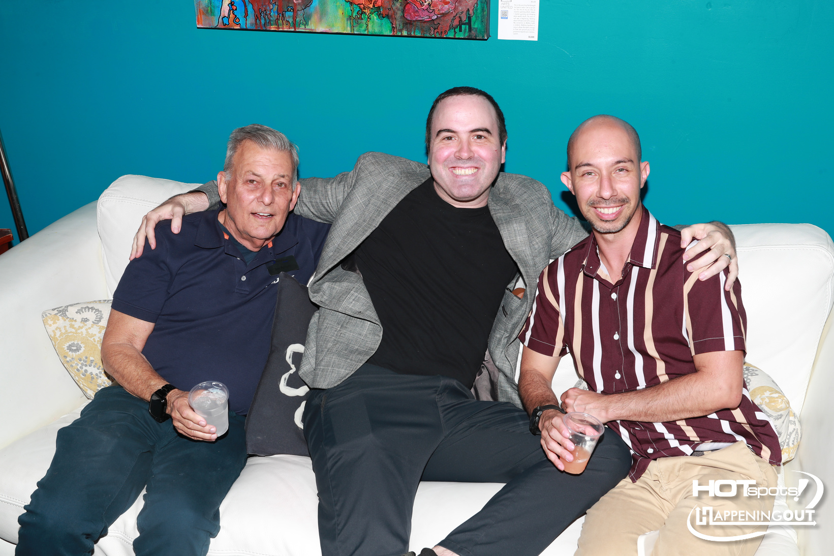 Three men sit closely on a white couch, smiling at the camera, each holding a drink in a casual setting with a teal wall behind them.