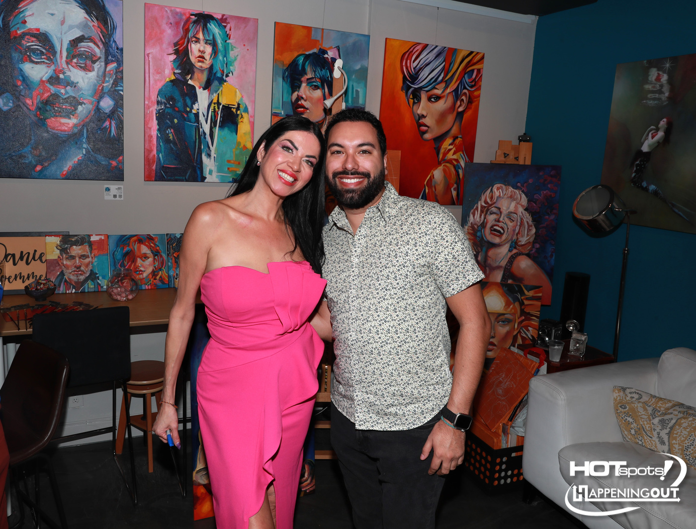 Smiling man and woman pose together in a colorful art studio with large vibrant paintings on the walls behind them.