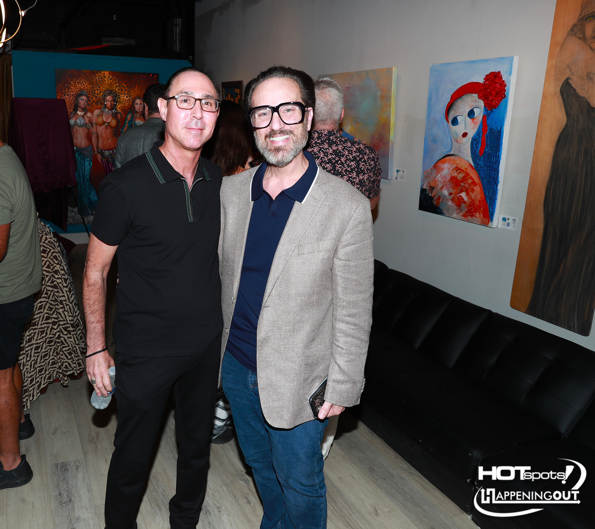 Two men posing at an art gallery, with colorful paintings on the wall and a black couch behind them in the background.
