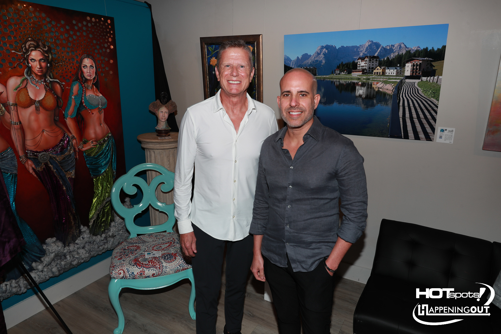 Two men pose together at an art gallery, with vibrant dancer paintings and a turquoise chair nearby.