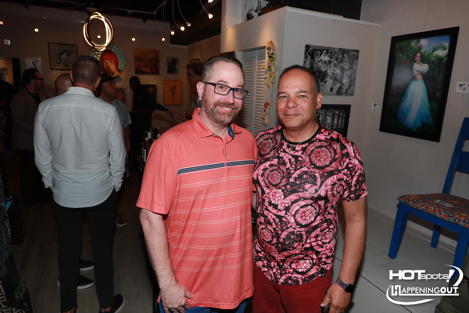 Two men pose for a photo at an art gallery, smiling with framed artworks on the wall behind them.