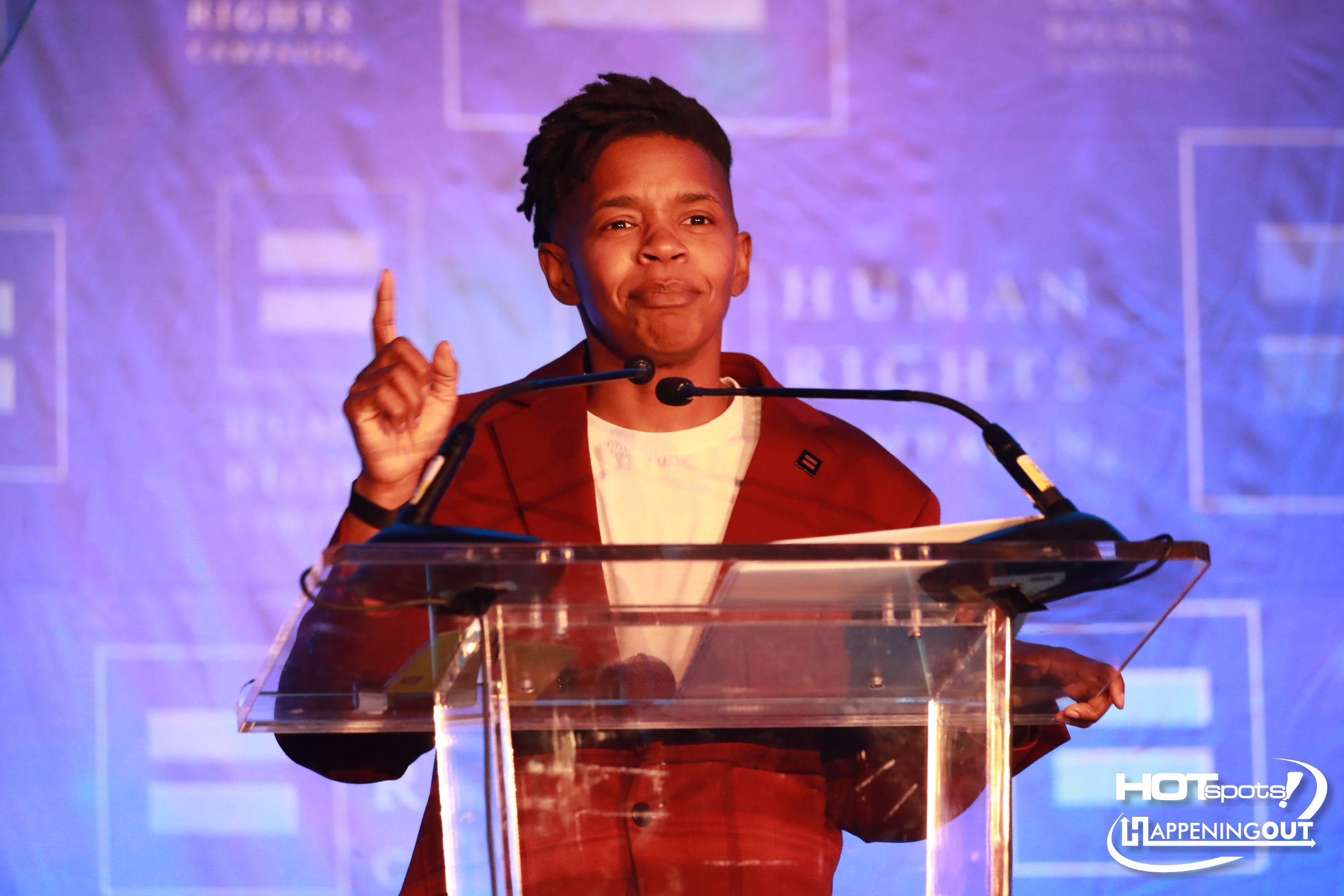 Male speaker in a red blazer at a clear podium on stage, raising one finger as he speaks into two microphones.