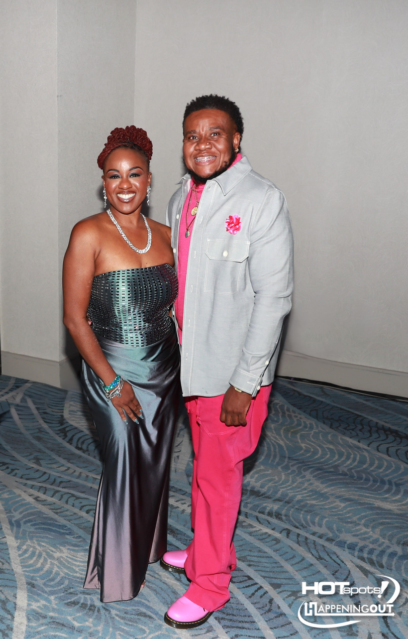 Smiling couple posing at a formal event; woman in a metallic strapless gown with jewelry, man in pink pants and a light gray jacket with a pink shirt.