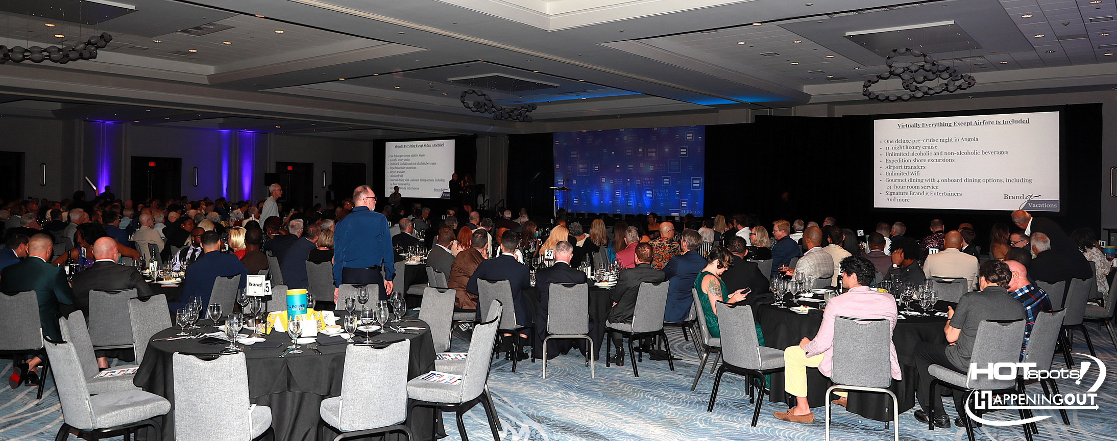Large conference hall filled with round tables and attendees watching a slide presentation on stage at the front. Two projection screens display content with a blue backdrop behind the speakers.