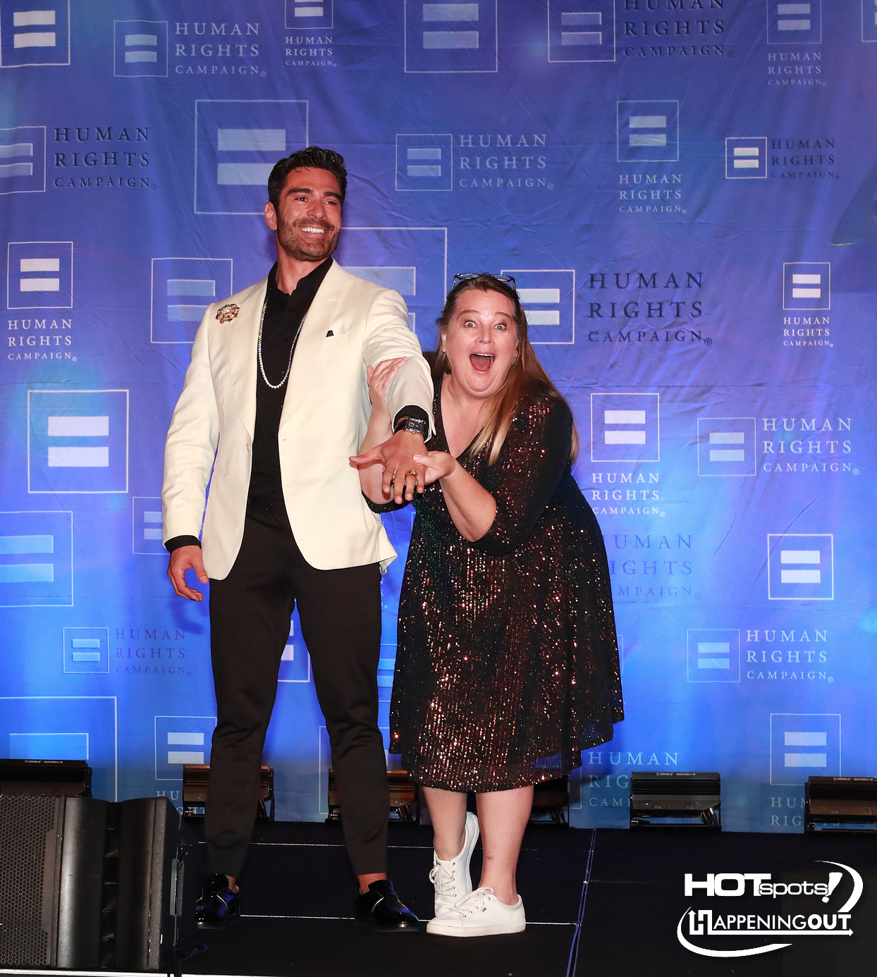 Man in a white blazer and a woman in a sequined dress pose together on stage at a Human Rights Campaign event.