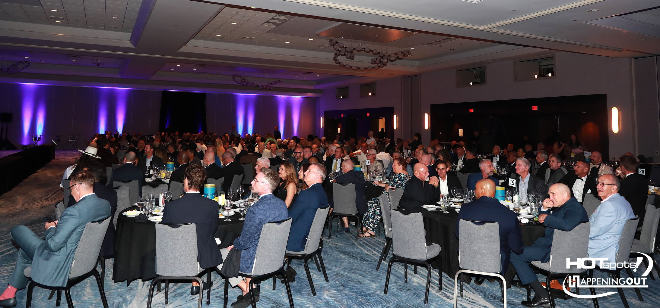 Large hotel ballroom filled with formally dressed guests seated at round tables for a banquet or gala, facing a stage with purple lighting awaiting an event.